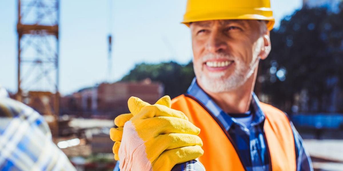 Baby Boomer construction worker in orange hat, gloves and vest shaking hands with a man in a blue plaid shirt on the job site