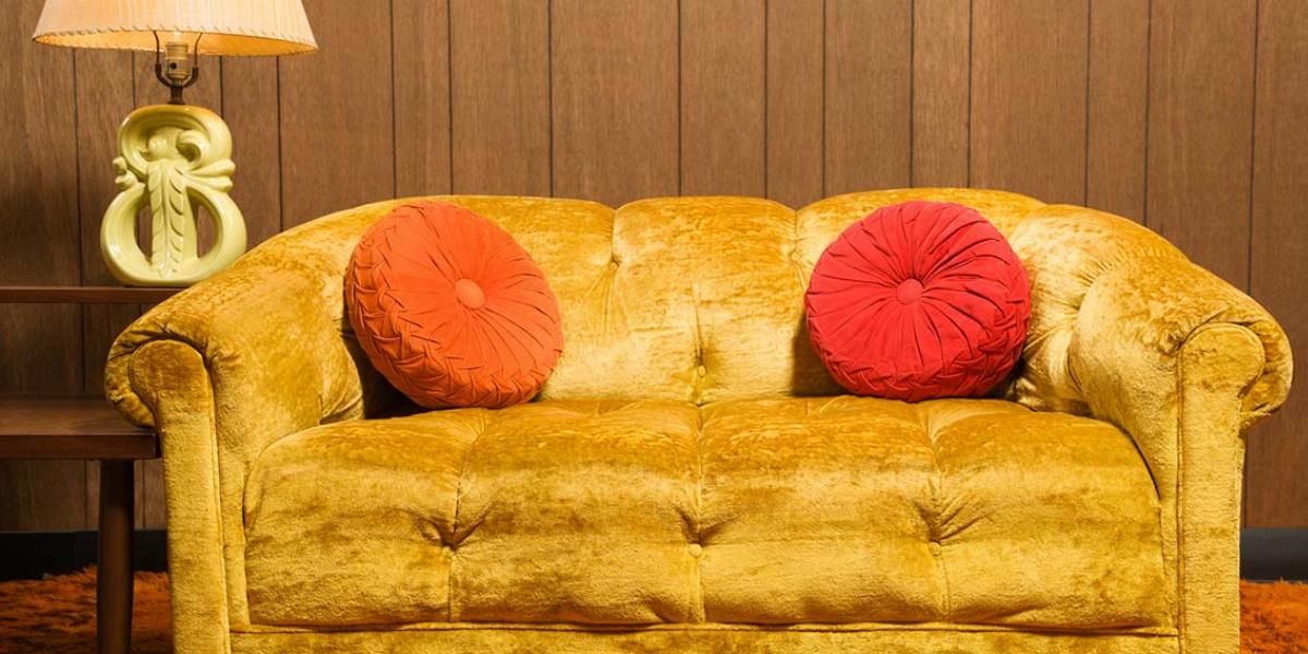 Yellow velvet sofa with red and orange pillows in front of 1970’s style wood paneling